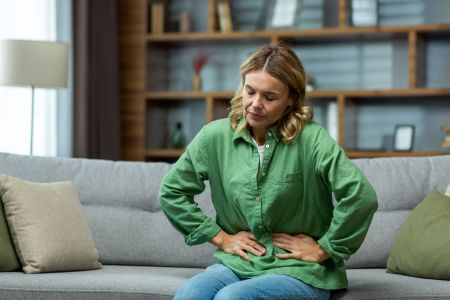 A woman sits on a sofa, wincing from abdominal discomfort